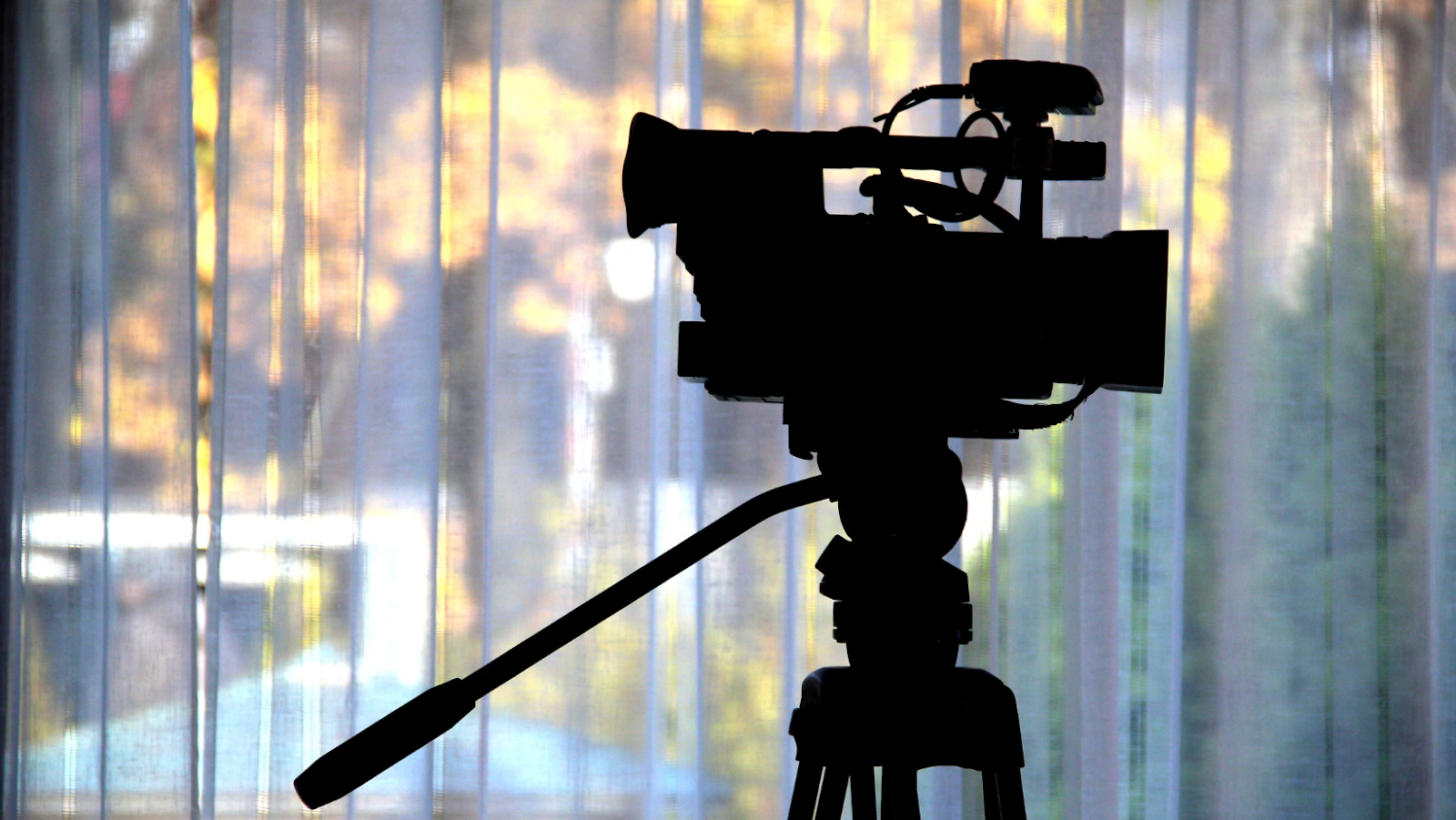 Silhouette of a professional video camera on a tripod in front of a window with vertical blinds.
