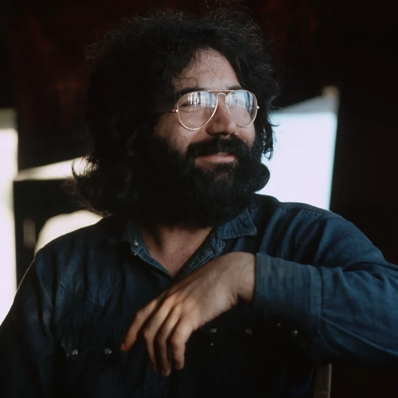 A man with curly hair, glasses, and a beard, wearing a denim shirt, sitting indoors with a relaxed expression.
